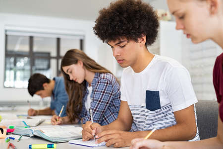Concentrated young man studying in classroom with friends sitting in a row. College student concentrating on studies while taking notes in university library. African american high school guy preparing exam with classmates in class.の写真素材