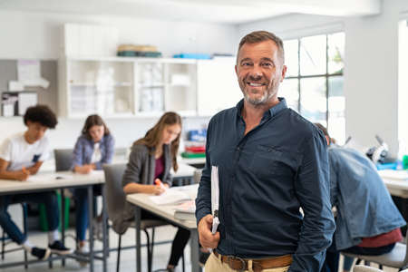 Portrait of mature teacher standing in university library and looking at camera with copy space. Happy mid adult lecturer at classroom standing after giving lecture. Satisfied high school teacher smiling and looking at camera while his studets studying in background.の写真素材