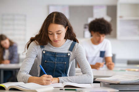High school student taking notes from book for her study. Young woman sitting at desk and finding information in college library. Focused girl studying in classroom completing assignment with classmate in background.の写真素材