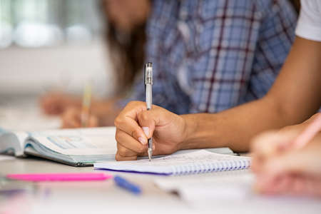 Close up of african student hand on table in classroom taking notes on notebook. Closeup of young man hand holding pen studying for exam in high school library. University student taking notes in book in classroom during lecture.の写真素材
