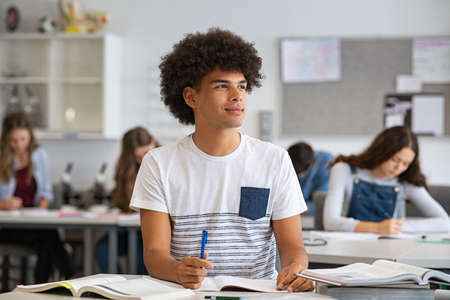 Portrait of contemplative young man during exam at high school, copy space. Smiling college student doing homework in class while looking away. Happy african young man preparing for exam in university classroom while thinking.の写真素材