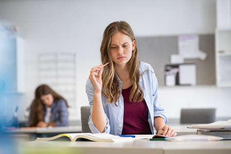 Stressed college student studying in the library at the university. Young woman having doubts while studying in high school classroom. Thoughtful girl thinking doing exam in class with copy space.の写真素材