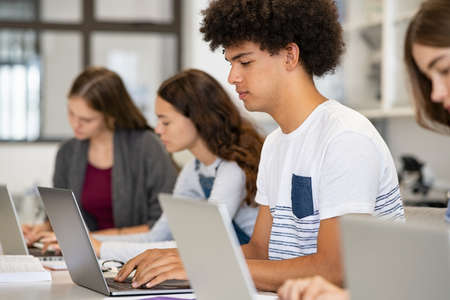 College student using laptop in class during computer lesson. Side view of guy sitting in a row studying in classroom on pc. Black high school student studying on laptop in university library.の写真素材