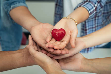 Close up of friends hands holding a red heart shape. Top view of young men and women hands showing small red heart. Multiethnic guys and girls holding a heart-shaped stone symbol of love and care.の写真素材