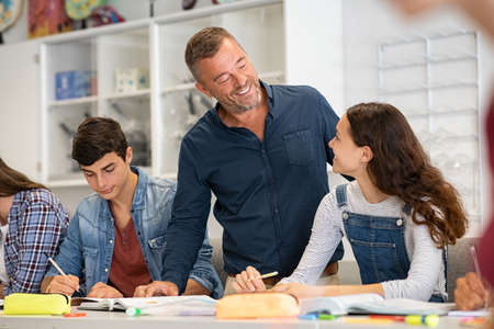 Happy mature professor helping student girl with her doubts while studying in class with friends. Smiling teacher in high school helping university student during exam. Group of young college classmates in classroom with helpful teacher during lecture.の写真素材