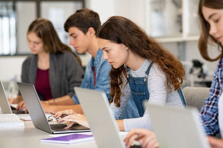 Serious college students studying on laptop sitting in a row in library. Young university multiethnic students using computer for study in classroom. Side view of casual girl typing on laptop during computer lesson.の写真素材