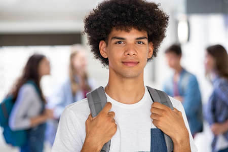 Portrait of university black student standing in hallway while looking at camera . Portrait of african guy holding school backpack with classmates talking in background. College young man in campus standing.の写真素材