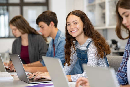 Happy young woman working on laptop and looking at camera in classroom. Portrait of smiling university student in library use computer for a research. Satisfied college student looking at camera while sitting in a row with classmates studying together.の写真素材