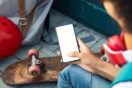 Close up of black guy hand using smartphone outdoor. High angle view of young man with skateboard and headphones using mobile phone with empty screen. Top view of teenager holding mobile phone with blank white screen.の写真素材