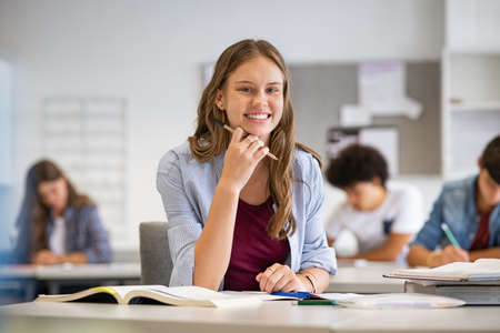 Portrait of smiling high school student with classmates in background writing notes in the classroom. Happy casual girl sitting at desk in class while looking at camera. Successful and smart young woman studying on book and notebook at college.の写真素材