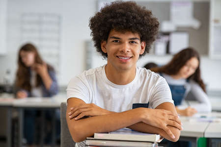 Portrait of happy african university student  in class looking at camera. Smiling young man at college leaning on stack of books at his desk with classmates in background. Satisfied african guy with hands on pile of books at college.の写真素材
