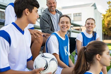 Young man player holding football sitting in stands with teammates. Smiling school girl talking to football player in school. Mixed members of high school soccer team talking about the strategy.の写真素材