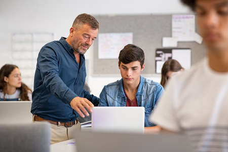 Professor assisting college student with laptop in classroom during computer lesson. Teacher talking and explaining something to guy sitting at desk. Mature man lecturer helping high school young man with laptop during lecture in university.の写真素材