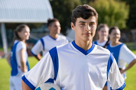 Portrait of young satisfied soccer player holding a football and looking at camera. Young man during training on soccer field with copy space. Proud teen guy training on soccer field with school teammmates in background.の写真素材