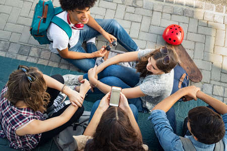 High angle view of guys and girls sitting and talking together. Cheerful group of happy friends in conversation after class. Top view of group of carefree teens spending time together after school while young woman using smart phone.の写真素材