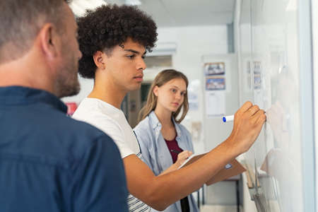 Young african american man solving mathematics problem on whiteboard with the help of his teacher. College student solving algebra equation on white board in classroom with students. High school black guy trying to understand mathematics problem during lesson with professor.の写真素材