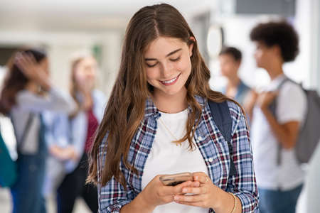 Young woman in high school campus using smartphone. Smiling university student texting on mobile phone standing in hallway. Happy college girl messaging on smart phone, using app.の写真素材