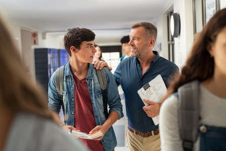Mature man talking to high school student in hallway at the end of the lesson. Senior professor in conversation with guy student in campus while walking at hallway. High school teacher holding tests and talking to encourage young man after exam.の写真素材