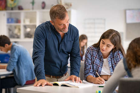 Secondary student being helped by professor during class. Mature man lecturer helping young woman during class test. High school girl in a lecture asking for explanations to helpful teacher.の写真素材