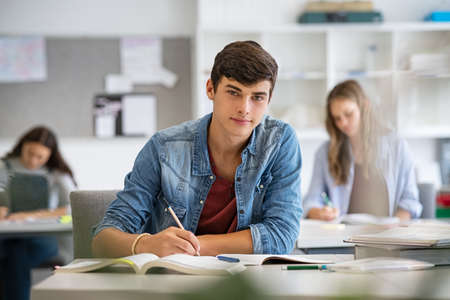Happy student taking notes while studying in high school. Satisfied young man looking at camera while sitting at desk in classroom. Portrait of college guy writing while completing project and assignment.の写真素材