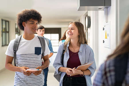 Multiethnic college friends walking in campus and talking while having break after classes. University students having fun after lesson at school. High school girl smiling while chatting with her black guy in hallway.の写真素材