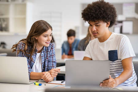 College students sitting together at desk and using laptop during computer lesson. Young black man and casual girl working on laptop in high school library. Happy smiling multiethnic friends working on laptop for an assignment.の写真素材