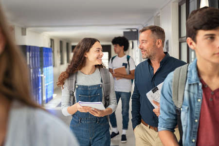Happy high school student talking to mature man professor in hallway. Senior man teacher in conversation to school girl holding book in campus. Helpful lecturer explaining concept to college student outside classroom.の写真素材