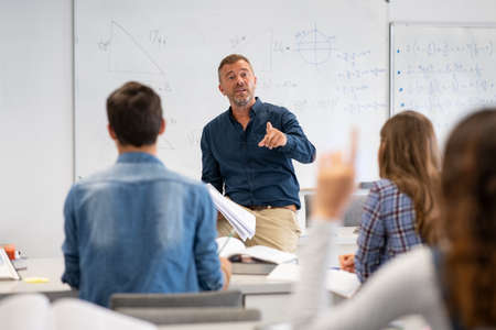 Professor pointing at college student with hand raised in classroom. Student raising a hand with a question for the teacher. Lecturer teaching in class while girl have a question to do during a math lesson.の写真素材