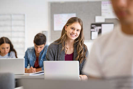 Happy college student studying using laptop in classroom during computer lesson. Portrait of beautiful and intelligent girl in library using laptop to study. High school student smiling and using computer while looking at camera.の写真素材