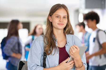 Portrait of happy satisfied girl standing in university hall with classmates in background. Pretty student holding digital tablet at high school and looking at camera. Smiling university young woman looking at camera after the end of the lessons.の写真素材