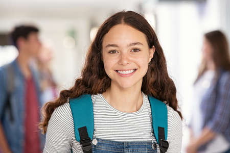 Portrait of beautiful college student standing in hallway school while looking at camera. Intelligent university girl with backpack smiling. Proud and satisfied young teen in high school corridor standing.の写真素材