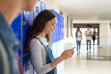 College student checking cellphone after class standing near locker with copy space. Happy smiling student reading message on smartphone while leaning on locker in college campus. University young casual woman messaging on smart phone standing in campus hallway.の写真素材