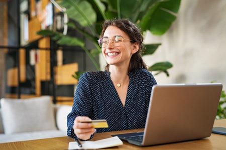 Young happy woman doing shopping online with her laptop at home. Portrait of excited woman holding credit card and buy on an e-commerce site with copy space. Beautiful laughing girl paying online bills using debit card and computer.の写真素材