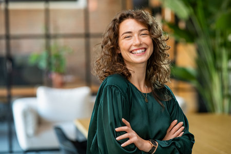 Portrait of young smiling woman looking at camera with crossed arms. Happy girl standing in creative office. Successful businesswoman standing in office with copy space.の写真素材