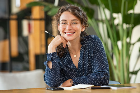 Portrait of smiling woman wearing spectacles and holding pencil while sitting at desk. Business woman taking notes in diary and looking at camera. University girl with eyeglasses sitting on table at library.の写真素材