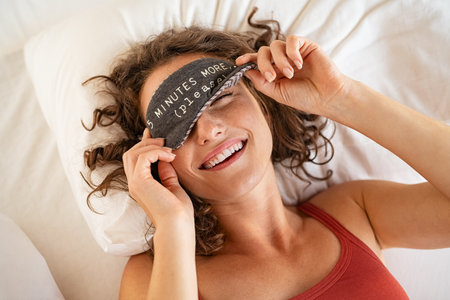 Top view of tired young woman sleeping on bed. Close up face of beautiful sleepy girl wearing funny eye mask while peeping with one eye. High angle view of woman covering her eyes with sleeping mask with five minutes more text.の写真素材