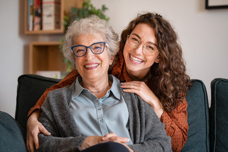 Portrait of old grandma and adult granddaughter hugging with love on sofa while looking at camera. Happy young woman with eyeglasses hugging from behind older grandma with spectacles. Senior woman spending time with her beautiful daughter, generation family concept.の写真素材