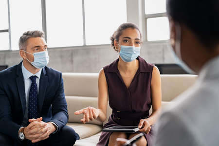 African businesswoman wearing face mask during pandemic sitting and discussing strategy with businesspeople in office. Group of multiethnic business team wearing protective mask and discussing work together during meeting. Young black woman and business man brainstorming new business plans during pandemic.の写真素材