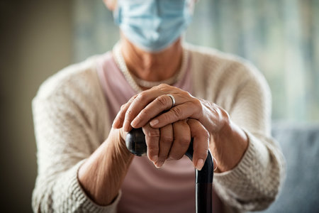 Close up of senior disabled woman hands holding walking stick and wearing face protective mask...