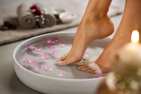 Close up of woman soaking her feet in grey bowl of water with floating petals at beauty luxury spa. Detail of woman feet at wellness center on pedicure procedure. Foot spa and body care concept.の写真素材