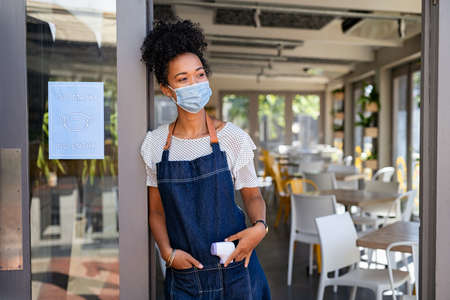 Portrait of happy young woman standing at doorway of her cafeteria while protective face mask during pandemic.の写真素材