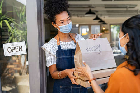 African restaurant owner working only with take away orders during outbreak. Young black woman wearing face mask giving takeout meal to customer outside her cafeteria.の写真素材