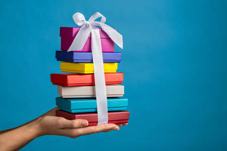 Woman holding colored boxes for gifts tied with satin ribbon. Girl holding pile of gifts boxes isolated against blue wall with copy space. Young woman with huge stack of presents for christmas or birthday.の写真素材