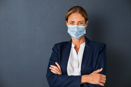 Portrait of happy businesswoman in formal clothing wearing face mask isolated against grey wall. Smiling entrepreneur wearing surgical mask looking at camera. Portrait of mature successful business woman with surgical face mask isolated on gray background with copy space.の写真素材