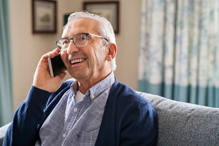 Smiling old man talking over smartphone while relaxing at home. Senior man after retirement using smart phone to connect with friends and family. Carefree elder talking over mobile phone with her daughter while sitting on couch and looking up.の写真素材