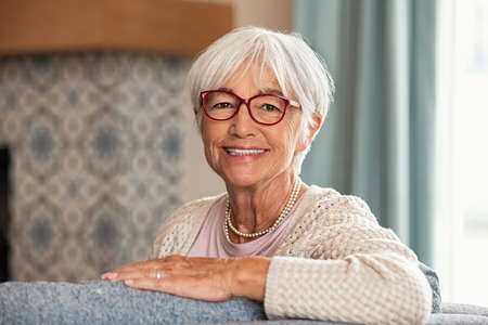 Portrait of beautiful senior woman with white hair and eyeglasses. Happy old woman relaxing on couch at home while looking at camera with big grin. Successful mature woman smiling while wearing red specs.の写真素材