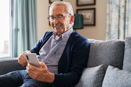 Smiling old man using smartphone while sitting on couch in living room. Happy elderly man with spectacles messaging on mobile phone at home. Healthy senior in video call with his family on smart phone while relaxing at nursing home.の写真素材