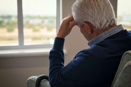 Rear view of sad senior man sitting near window at home. Tired and depressed old man sitting on armchair in living room feeling hurt and lonely. Frustrated senior sitting on sofa at nursing home with head leaning on his hand while looking through the window.の写真素材