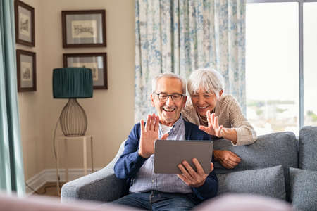 Happy senior couple using digital tablet for video conference and waving hand while laughing at home. Smiling grandparents making video call and greeting the family sitting on sofa. Old man and cheerful woman using digital tablet for video call with nephews or friends.の写真素材