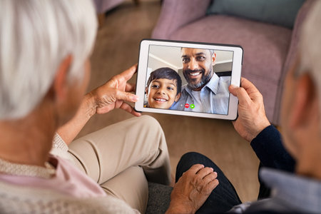 Rear view of senior couple doing a video call with son and grandson during covid19 pandemic. Close up of hands of old grandparents holding digital tablet while in conversation with little grandson. Happy indian father with child greeting in video conference his parents.の写真素材